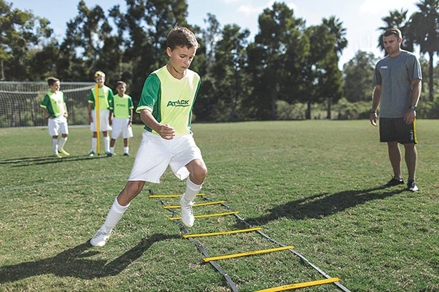 Boy performing agility drill on yellow and black speed ladder on soccer field with coach and teammates in background