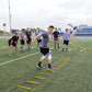 Athlete performing agility ladder drills on artificial turf field with training group watching in background