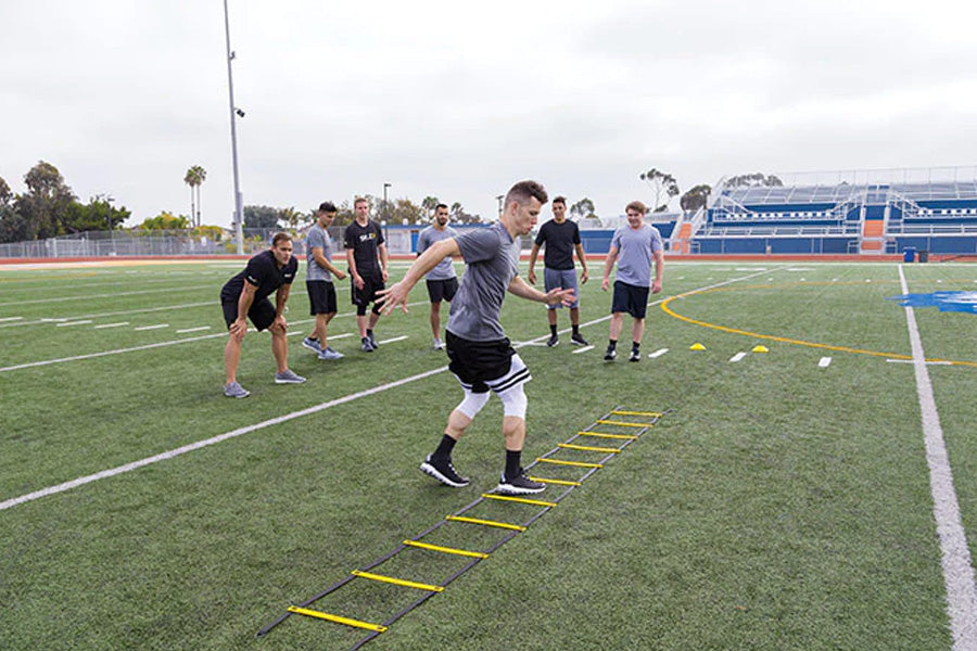 Athlete performing agility ladder drills on artificial turf field with training group watching in background
