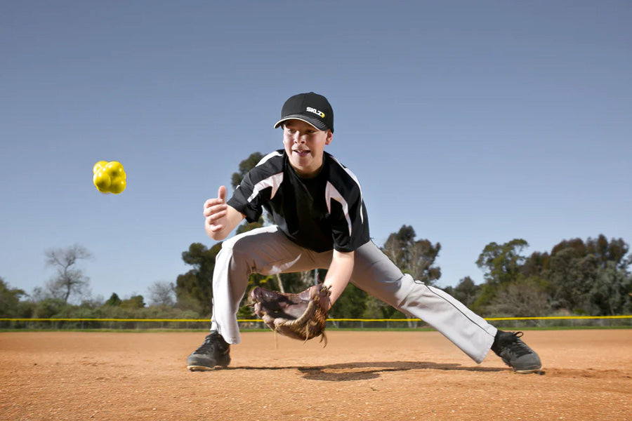 SKLZ yellow reaction training ball in mid-air above baseball player in fielding position on dirt field