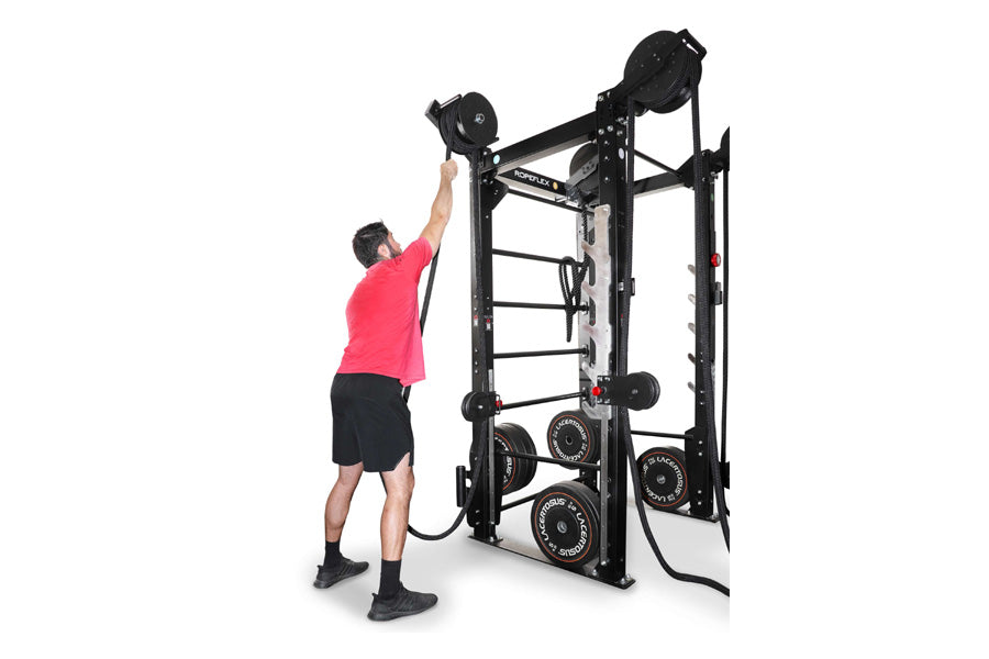 Man in pink shirt performing overhead dumbbell press on black Ropeflex RX2100 functional trainer with weight plates