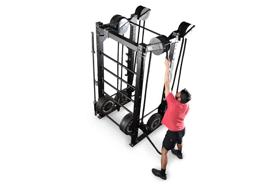 Black steel cable machine with dual pulleys and weighted stack, man in pink shirt demonstrating overhead pull exercise