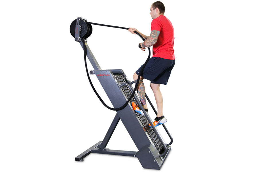 Man in red shirt and navy shorts using black and gray rope climbing machine with resistance wheel against white background