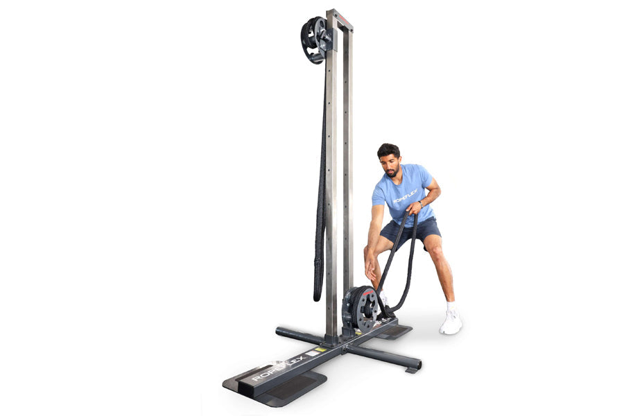 Man in blue shirt and navy shorts using tall vertical rope training machine with pulley system on white background
