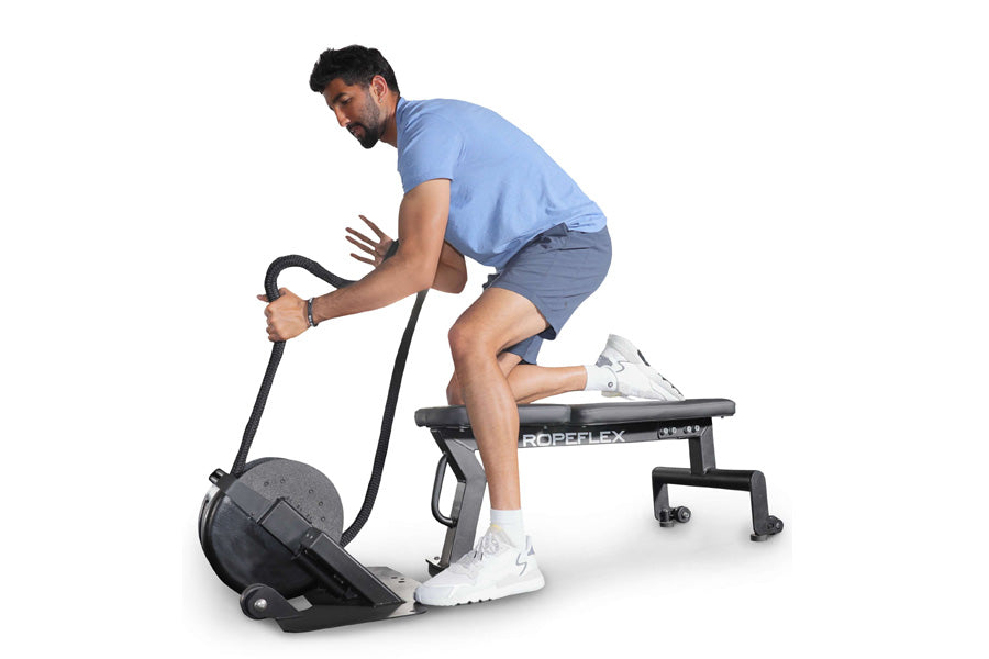 Man using black Ropeflex RX2000 rope trainer while seated on exercise bench, wearing light blue shirt and navy shorts