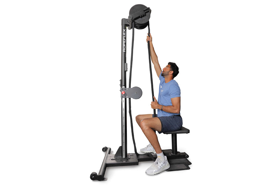 Man using black Ropeflex RX2500 upright rope trainer with pulley system and weighted ball, seated on attached bench