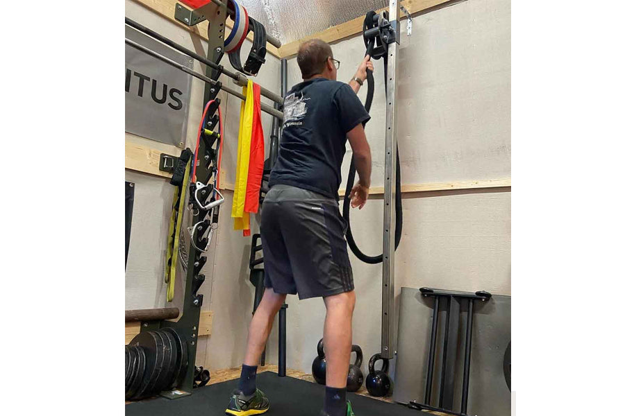 Man wearing black t-shirt and shorts using rope drum attachment mounted on wall in gym setting