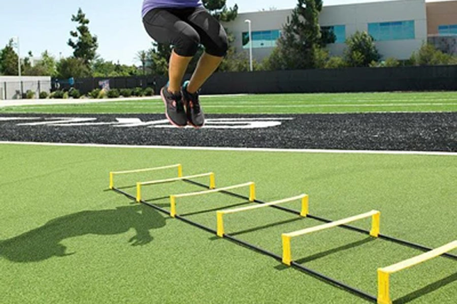 Athlete jumping over yellow and black agility ladder on artificial turf field during training