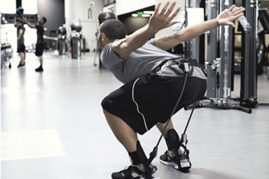 Man wearing gray t-shirt and black shorts with white stripe performing squat exercise with black SKLZ Hopz resistance bands i