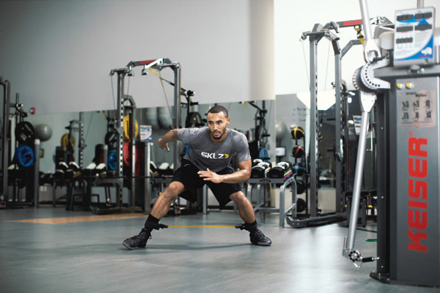 Man performing lateral lunge with SKLZ Lateral Resistor Pro resistance band in gym setting surrounded by exercise equipment