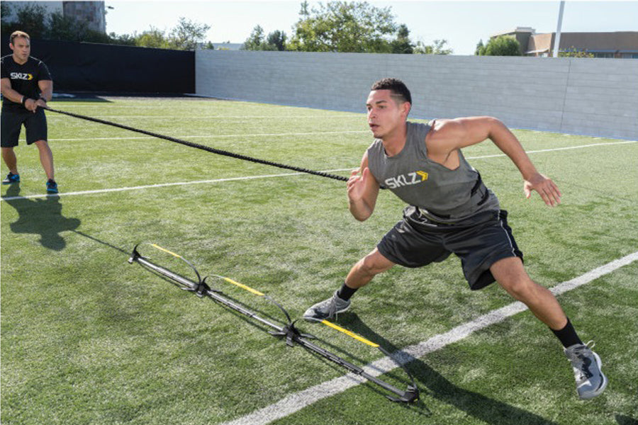 Male athlete performing sled push drill on grass field wearing gray tank top and black shorts with resistance harness attache