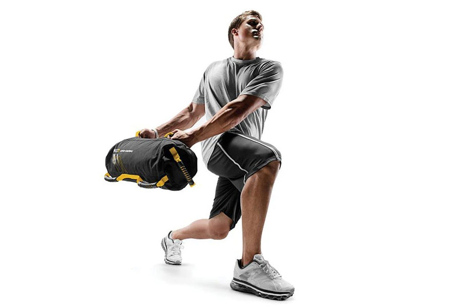 Man wearing gray t-shirt and black shorts holding black and yellow weighted sandbag while exercising on white background