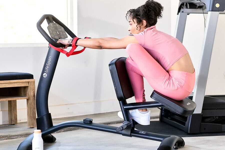 Woman in pink athletic wear using stationary stretch machine with digital display in home gym setting