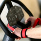 Hands gripping black exercise machine handlebar with red wrist straps and digital display panel visible