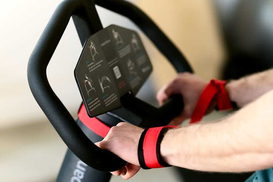 Hands gripping black exercise machine handlebar with red wrist straps and digital display panel visible