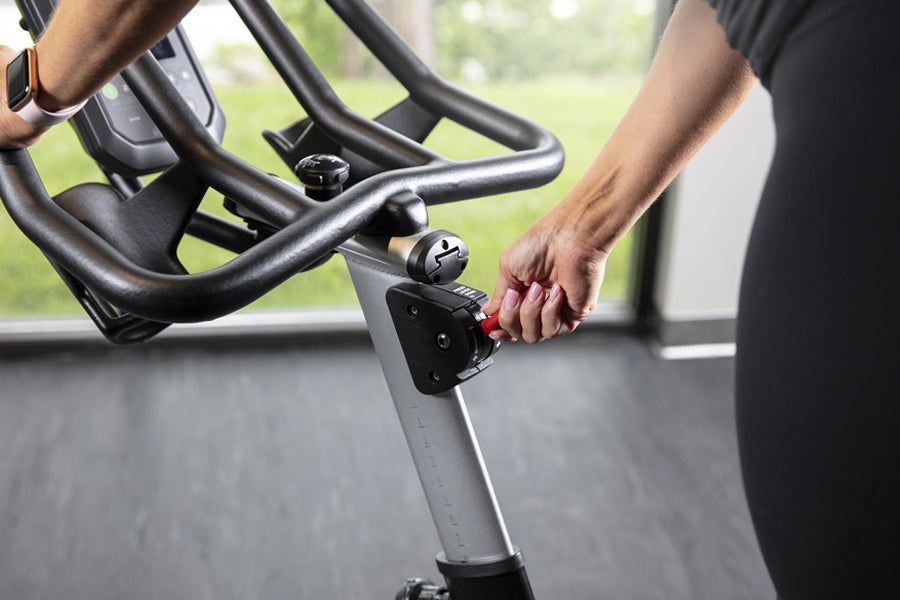 Person adjusting red resistance knob on black and gray stationary exercise bike with curved handlebars