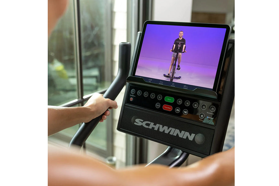 Person holding handlebar of Schwinn 190 upright exercise bike displaying screen with cyclist on purple background