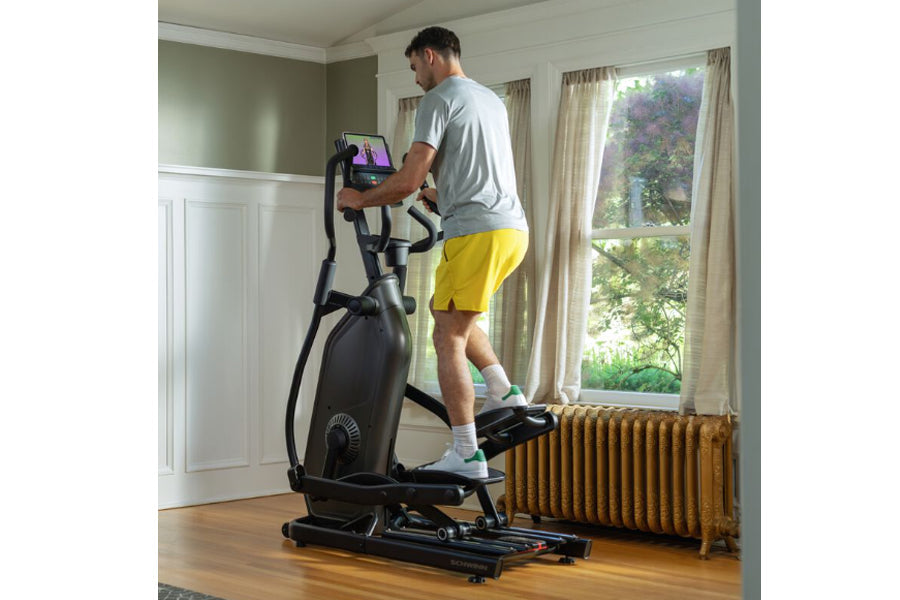 Man wearing gray shirt and yellow shorts using black Schwinn 490 elliptical machine in home gym with hardwood floor