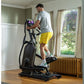 Man in gray shirt and yellow shorts using black elliptical machine with digital display in home gym setting