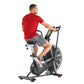 Man wearing red t-shirt and gray shorts riding black and red Schwinn Airdyne stationary exercise bike on white background