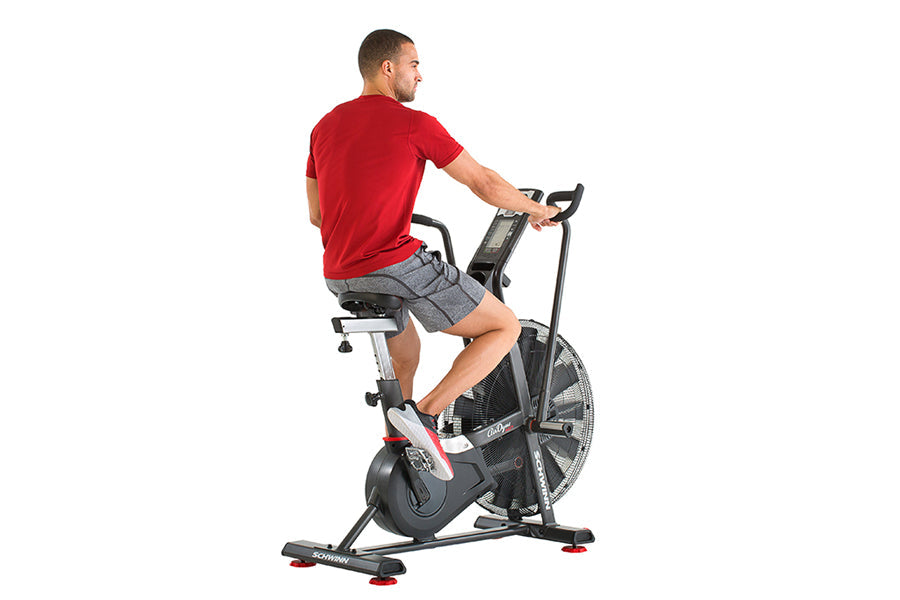 Man wearing red t-shirt and gray shorts riding black and red Schwinn Airdyne stationary exercise bike on white background