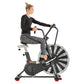 Woman wearing black long-sleeve top and gray patterned leggings riding Schwinn Airdyne AD7 exercise bike with large fan wheel