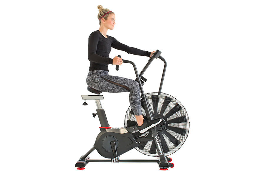 Woman wearing black long-sleeve top and gray patterned leggings riding Schwinn Airdyne AD7 exercise bike with large fan wheel