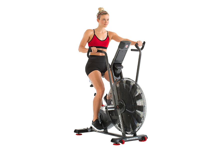 Woman wearing red sports bra and black shorts using black Schwinn Airdyne stationary exercise bike with large fan wheel on wh