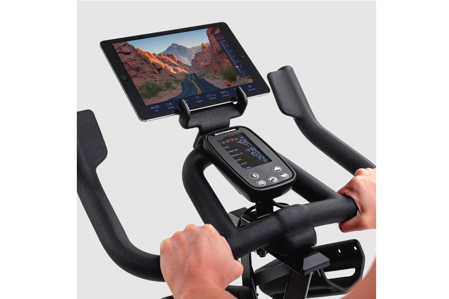 Black upright stationary bike with foam-padded handlebars, digital console, and tablet holder displaying fitness app, worn by