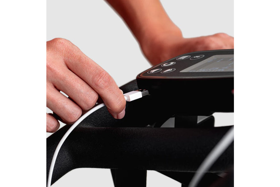 Hand plugging charging cable into black indoor exercise bike with digital display