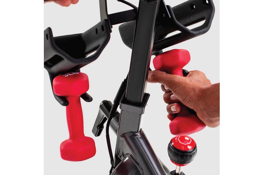 Schwinn IC4 indoor cycling bike with black frame, red handlebar grips, and red dumbbell weights held by model's hands
