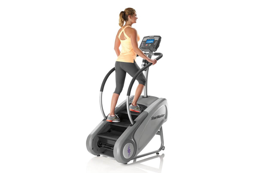 Woman using gray StairMaster SM3 StepMill cardio machine with digital display, wearing yellow tank top and black capris