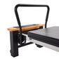 Black and wood Pilates reformer with padded black platform, arched handle bar, and adjustable foot bar on white background