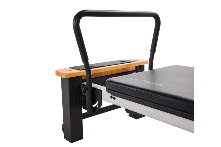 Black and wood Pilates reformer with padded black platform, arched handle bar, and adjustable foot bar on white background