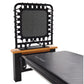 Black and gray Pilates reformer with wood armrests and black elastic cords attached to headrest on white background