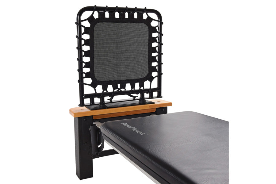 Black and gray Pilates reformer with wood armrests and black elastic cords attached to headrest on white background