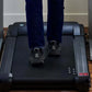 Person wearing dark jeans and black athletic shoes walking on black under-desk treadmill with silver handles on wooden floor