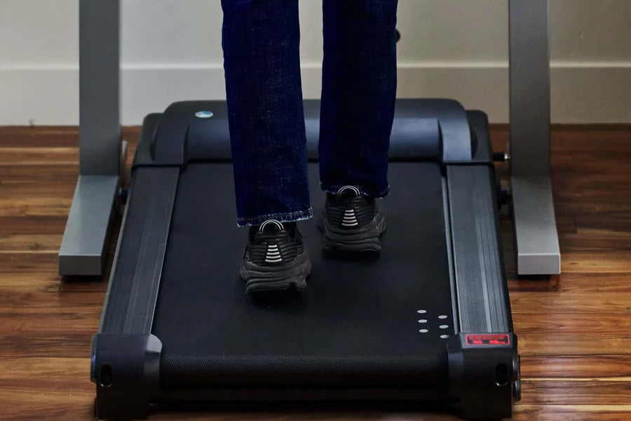 Person wearing dark jeans and black athletic shoes walking on black under-desk treadmill with silver handles on wooden floor