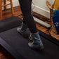 Black under-desk treadmill with cushioned surface, worn by model in black socks and boots on wooden floor