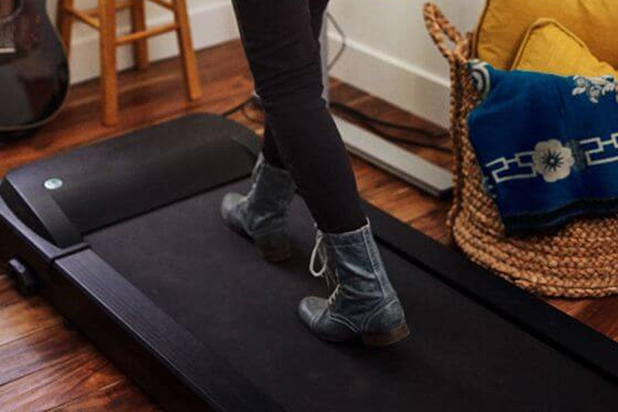 Black under-desk treadmill with cushioned surface, worn by model in black socks and boots on wooden floor