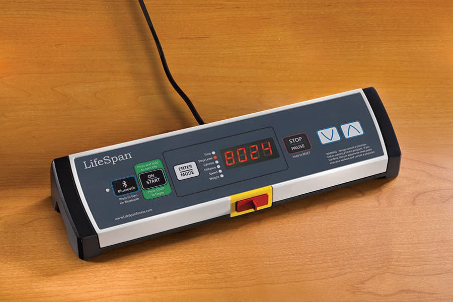 LifeSpan TR1000-GlowUp under-desk treadmill with digital display, control buttons, and yellow emergency stop switch on wood s