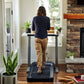 Woman in navy pullover and tan pants using black LifeSpan treadmill desk in home office with brick fireplace