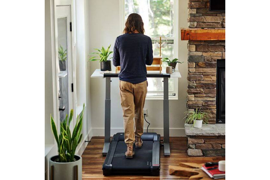 Woman in navy pullover and tan pants using black LifeSpan treadmill desk in home office with brick fireplace