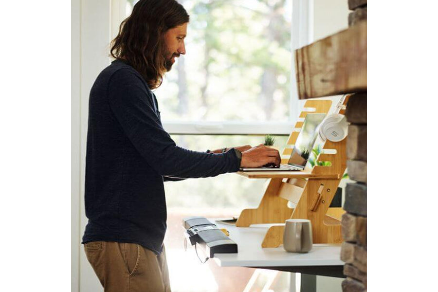 Man with long dark hair wearing navy blue long-sleeve shirt and tan pants using laptop at wooden standing desk organizer with