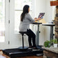 Woman in gray top and black pants using laptop while standing on black treadmill desk with adjustable height platform