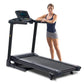 Black folding treadmill with digital display console, shown with woman in navy tank top and gray shorts standing beside it on