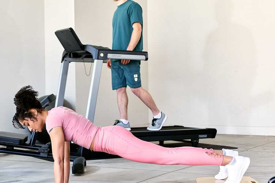 Woman in pink workout set doing plank exercise on floor while man in teal shirt runs on LifeSpan TR3000i folding treadmill in