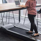 Person in red patterned top walking on black under-desk treadmill while working at wooden table in modern office
