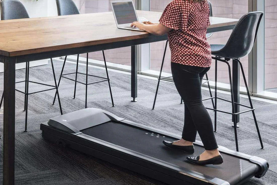 Person in red patterned top walking on black under-desk treadmill while working at wooden table in modern office