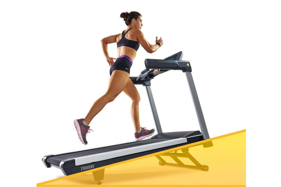 Woman running on LifeSpan TR6000i treadmill with black and gray frame, wearing navy sports bra and purple shorts against whit